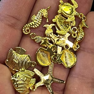 May include: A collection of gold-colored charms in various sea life shapes, including seahorses, fish, starfish, and shells. Each charm has a small loop for attaching to jewelry or other crafts. The charms are highly detailed and have a shiny finish.