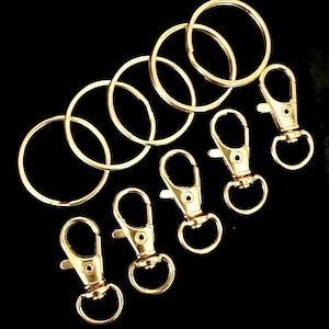 May include: Gold-colored key rings and lobster claw clasps arranged on a black background. The key rings are circular, and the clasps have a swivel design. These hardware components are suitable for crafting and jewelry making.
