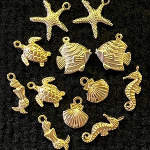 May include: A collection of gold-colored charms in various sea-themed shapes, including starfish, fish, turtles, mermaids, seahorses, and seashells. Each charm has a small loop for attaching to jewelry or other crafts. The charms are arranged on a dark background.