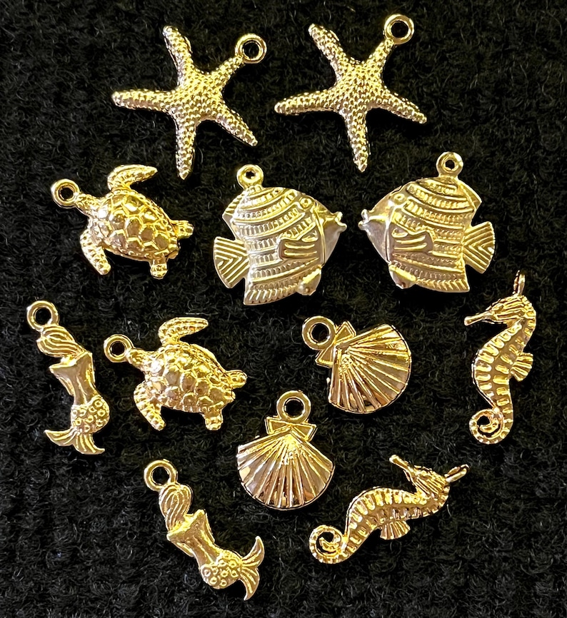 May include: A collection of gold-colored charms in various sea-themed shapes, including starfish, fish, turtles, mermaids, seahorses, and seashells. Each charm has a small loop for attaching to jewelry or other crafts. The charms are arranged on a dark background.