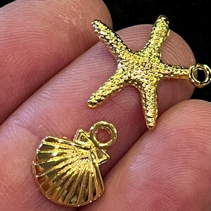 May include: Two gold-colored charms: a starfish and a seashell. The starfish has a textured surface and five points. The seashell charm has a fan-like design with a loop for attaching to jewelry. Both charms are shiny.
