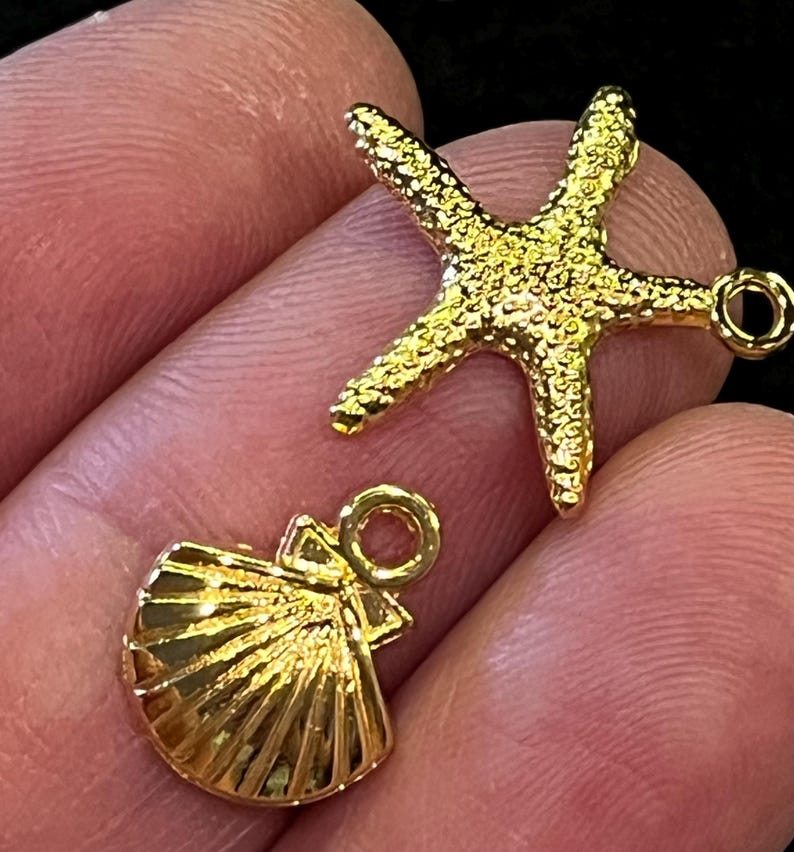May include: Two gold-colored charms: a starfish and a seashell. The starfish has a textured surface and five points. The seashell charm has a fan-like design with a loop for attaching to jewelry. Both charms are shiny.