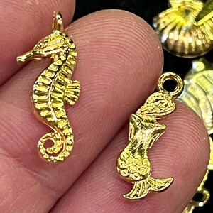 May include: Two gold-colored charms. One is a seahorse, detailed with ridges and fins. The other is a mermaid with a flowing tail and detailed scales. Both charms have a loop at the top for attaching to jewelry.
