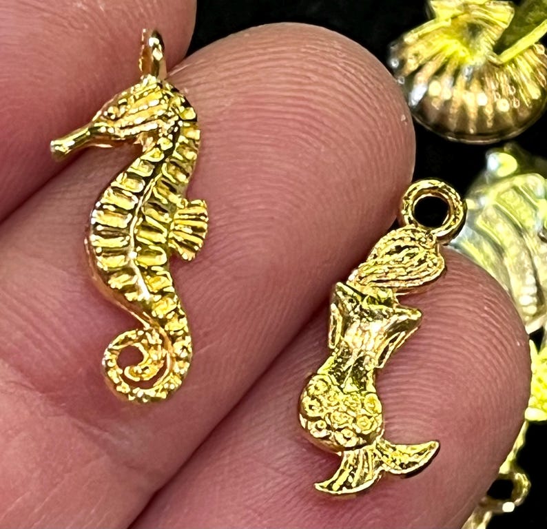 May include: Two gold-colored charms. One is a seahorse, detailed with ridges and fins. The other is a mermaid with a flowing tail and detailed scales. Both charms have a loop at the top for attaching to jewelry.