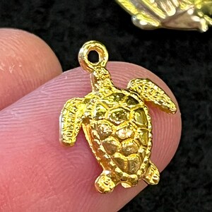 May include: A small, gold-colored turtle charm with detailed shell and flipper designs. The charm has a loop at the top for attaching to a necklace or bracelet. The turtle is held between two fingers against a dark background.