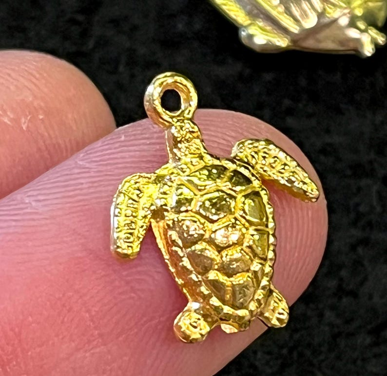 May include: A small, gold-colored turtle charm with detailed shell and flipper designs. The charm has a loop at the top for attaching to a necklace or bracelet. The turtle is held between two fingers against a dark background.