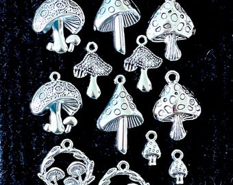 Könnte beinhalten: Eine Sammlung silberfarbener Pilzanhänger in verschiedenen Größen und Designs. Einige Anhänger haben detaillierte Kappen mit Mustern, andere ein glattes, gepunktetes Aussehen. Die Anhänger sind auf einer dunklen, strukturierten Oberfläche angeordnet, ideal für Schmuckherstellung oder Bastelarbeiten.