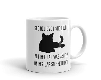 May include: White ceramic coffee mug with a black silhouette of a cat curled up on a lap. The mug has the text "She believed she could but her cat was asleep on her lap so she didn't" printed on it.