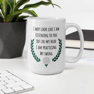 May include: White ceramic coffee mug with a green laurel wreath design and a golf ball graphic. The mug has black text that reads "I may look like I am listening to you but in my head I am practicing my swing."