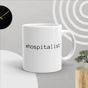 Può includere: Tazza da caffè in ceramica bianca con un hashtag nero e il testo "#hospitalist" stampato sul davanti.