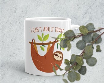 May include: White ceramic mug featuring a brown sloth hanging from a branch with green leaves. The mug has the text "I can't adult today" printed on it.