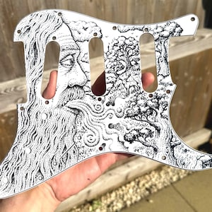 May include: A white guitar pickguard with a black and white illustration. The design depicts a bearded man's profile and a landscape with trees and swirling patterns. The pickguard has pre-drilled holes for mounting on a guitar.