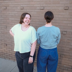 May include: Two people wearing casual clothing stand in front of a brick wall. One person wears a light green t-shirt and dark gray pants. The other person wears a blue sweatshirt and blue jeans with heart-shaped pockets.