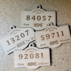 May include: Four wooden Christmas ornaments with engraved text. Each ornament has a rectangular shape with a rounded top and a hole for hanging. The ornaments display the year "2022", a zip code, and the word "HOME" along with a city and state.