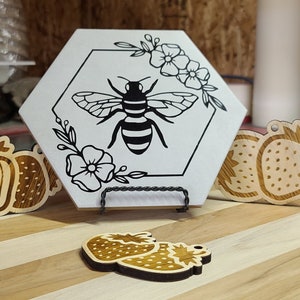 May include: A white hexagonal tile with a black and white bee illustration, flowers, and leaves. The tile is on a wooden stand. There are three wooden strawberry ornaments on a wooden surface.
