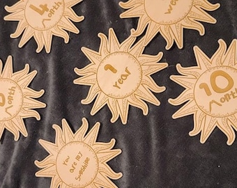 May include: A set of ten wooden sun-shaped baby milestone cards. The cards feature gold text with the phrases "1 month", "4 month", "6 month", "1 year", "10 month", and "You are my sunshine".