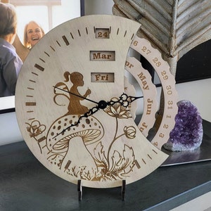 May include: Wooden clock with a whimsical design featuring a mushroom, a fairy, and floral accents. The clock face has a calendar with the months of March, May, and June. The clock hands are black and the clock face is white.