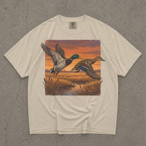 May include: Cream-colored t-shirt featuring a rectangular graphic of two ducks in flight over a marsh at sunset. The ducks have detailed plumage, with one displaying a green head. The shirt has a crew neck and short sleeves.
