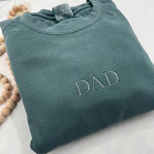 May include: A teal blue sweatshirt with the word "DAD" embroidered in white on the front.