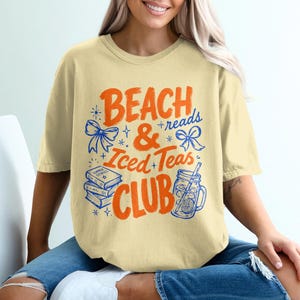 Può includere: T-shirt giallo chiaro con la scritta "Beach & Reads + Iced Teas Club" in arancione e blu. Il design include illustrazioni di libri, fiocchi e un barattolo con cannuccia.