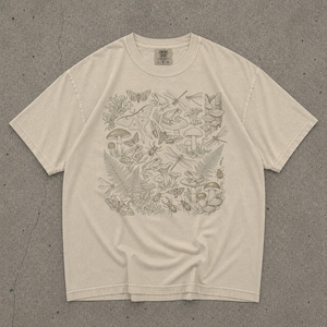 May include: Cream-colored t-shirt featuring a detailed illustration of various forest elements. The design includes mushrooms, insects, frogs, ferns, and butterflies. The shirt has a classic crew neck and short sleeves, with a Comfort Colors tag.