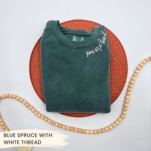 May include: A dark green sweatshirt with the text "you are loved" embroidered in white thread on the neckline.
