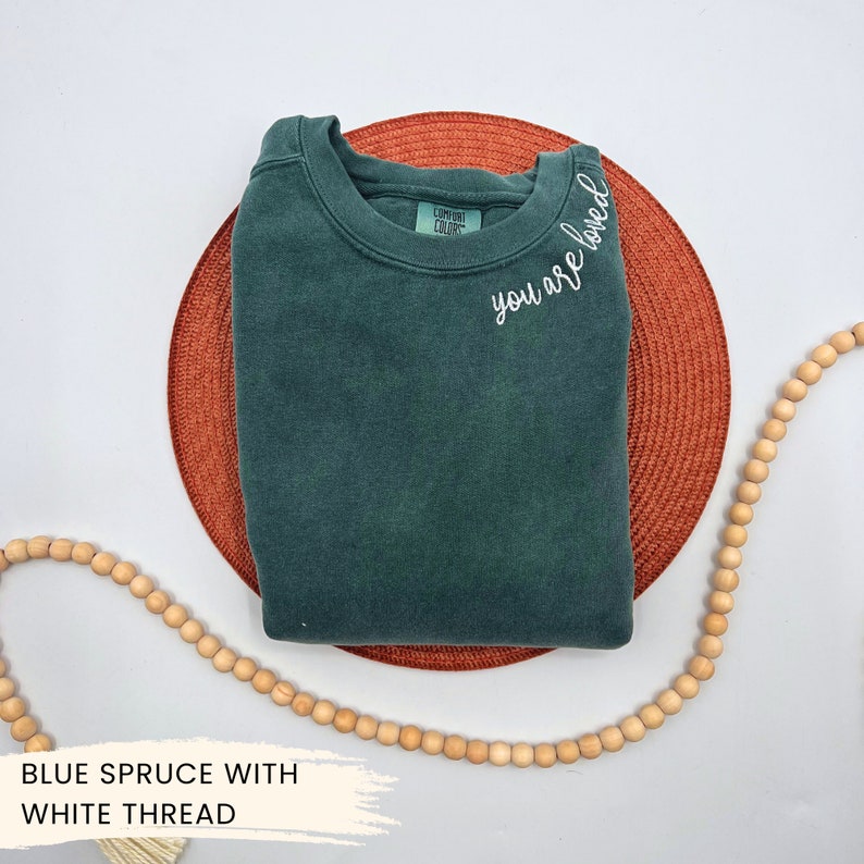 May include: A dark green sweatshirt with the text "you are loved" embroidered in white thread on the neckline.