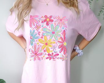 Puede incluir: Una camiseta rosa con un estampado floral colorido. Las flores están dibujadas en un estilo caprichoso con colores rosa, azul, amarillo y verde.