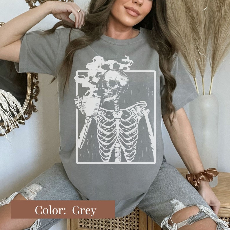 May include: A grey t-shirt featuring a white graphic of a skeleton holding a coffee cup, with steam rising from the cup. The skeleton is inside a white rectangular frame. The shirt is a solid grey color.