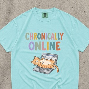 May include: A light turquoise short-sleeve t-shirt featuring the text "CHRONICALLY ONLINE" in multi-coloured letters. Below the text is a graphic of an orange tabby cat lying on its back on top of an open silver laptop computer.