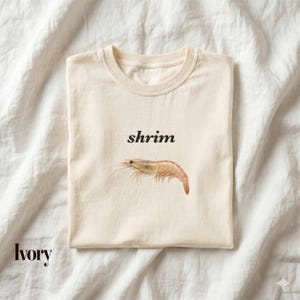 May include: An ivory-coloured short-sleeve t-shirt featuring the black text "shrim" printed above a realistic illustration of a single shrimp. The garment has a crew neckline and is displayed flat, showcasing the graphic centred on the chest.