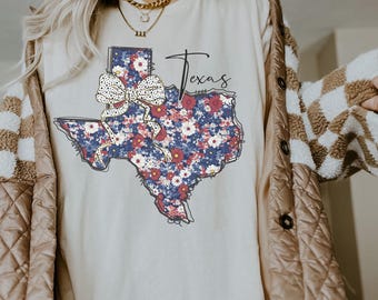 May include: A white t-shirt featuring a floral print of the state of Texas. The print includes a large bow in the centre of the state. The text "Texas" is written in a cursive font above the state.