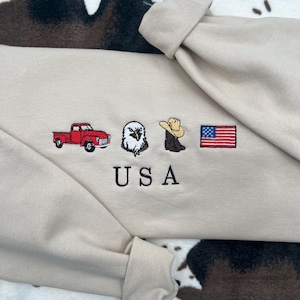 May include: A beige sweatshirt with embroidered images of a red pickup truck, an eagle, a cowboy hat, and an American flag. The word "USA" is embroidered below the images.