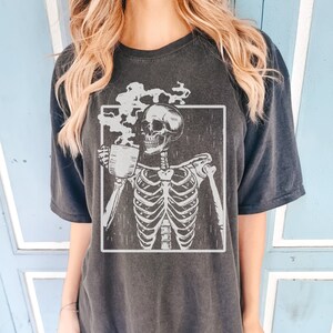 May include: Dark grey t-shirt featuring a skeleton holding a coffee cup, with steam rising from the cup. The skeleton is inside a white rectangular frame. The shirt has short sleeves and a relaxed fit.