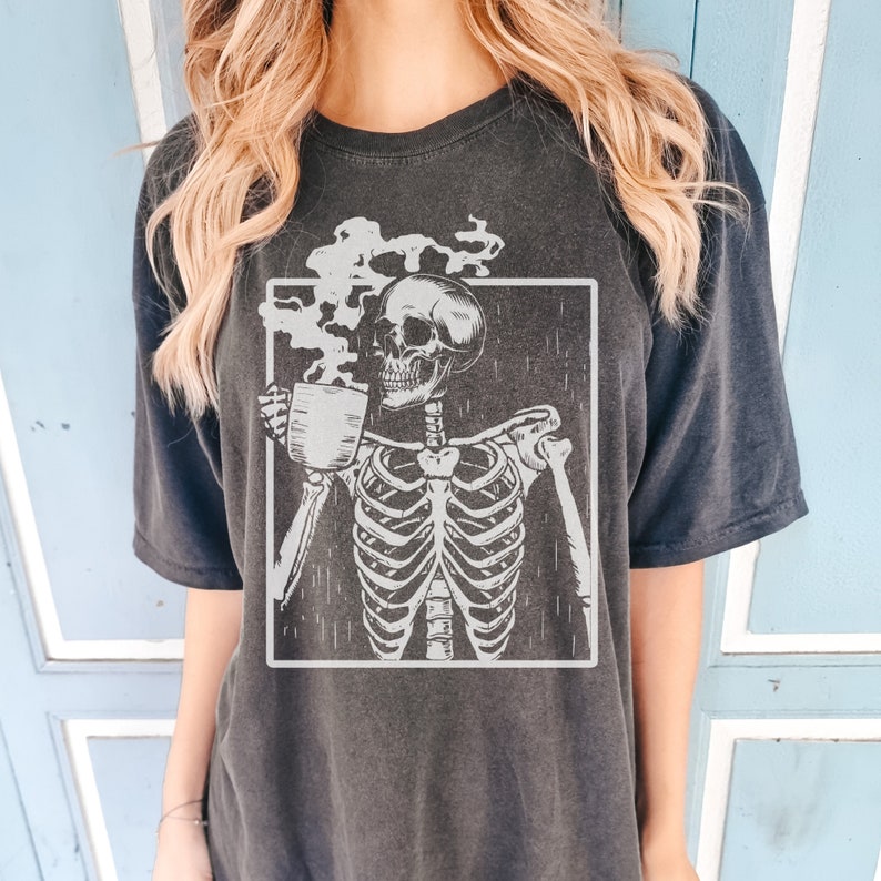 May include: Dark grey t-shirt featuring a skeleton holding a coffee cup, with steam rising from the cup. The skeleton is inside a white rectangular frame. The shirt has short sleeves and a relaxed fit.