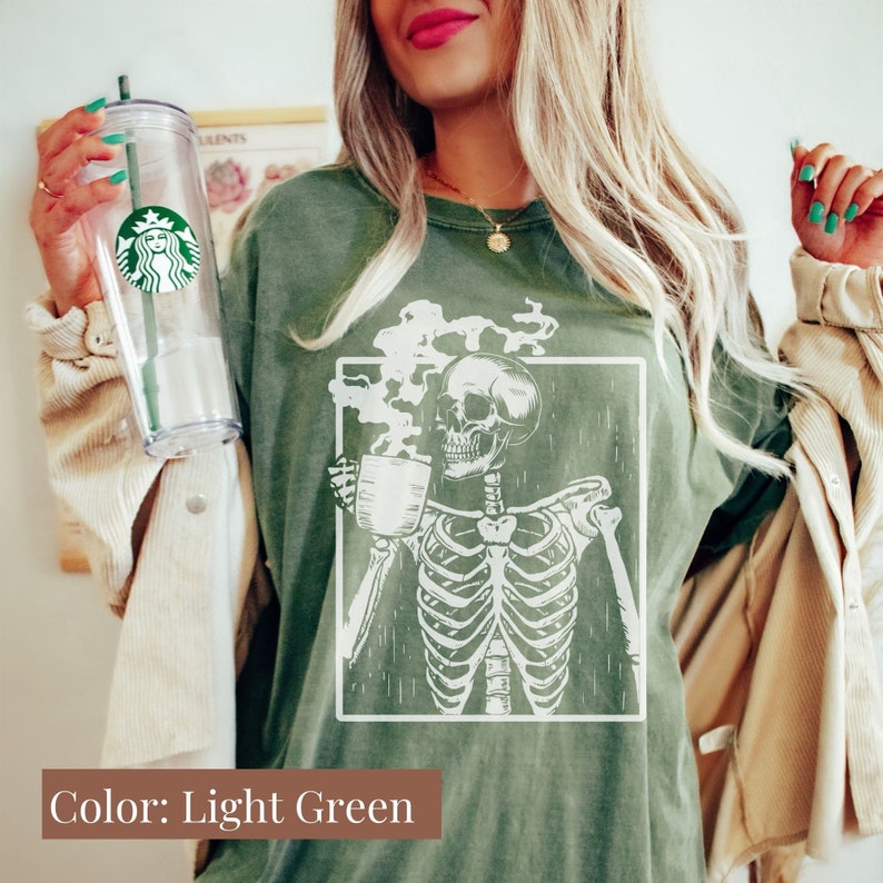 May include: Light green t-shirt featuring a white graphic of a skeleton holding a coffee cup, with steam rising. The text "Color: Light Green" is at the bottom. A clear Starbucks tumbler with a green straw is held in the image.
