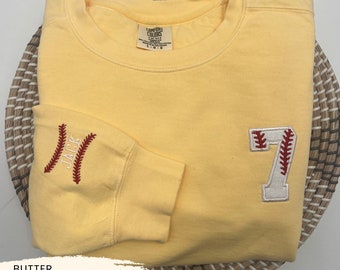 May include: A yellow sweatshirt with a white baseball number 7 with red stitching on the right side and the name "JACK" with red stitching on the left sleeve. The sweatshirt is on a woven basket.
