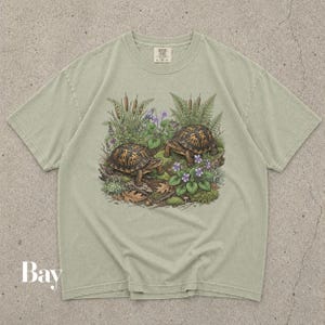 Op de afbeelding: Een saliegroen t-shirt met korte mouwen en een gedetailleerde afbeelding van twee doosschildpadden omringd door lisdodden, varens, mos en paarse bloemen. Het shirt heeft een ronde hals en een Comfort Colors-label in de binnenkraag.