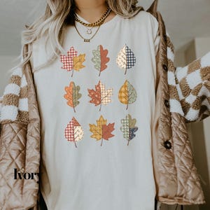 Könnte beinhalten: Ein elfenbeinfarbenes T-Shirt mit einem Grafikdruck von neun Herbstblättern, die in drei Reihen angeordnet sind. Jedes Blatt zeigt ein einzigartiges Patchwork-Design mit verschiedenen Mustern wie Karos, Vichy-Karo und Blumenprints in Rot-, Orange-, Gelb- und Blautönen.