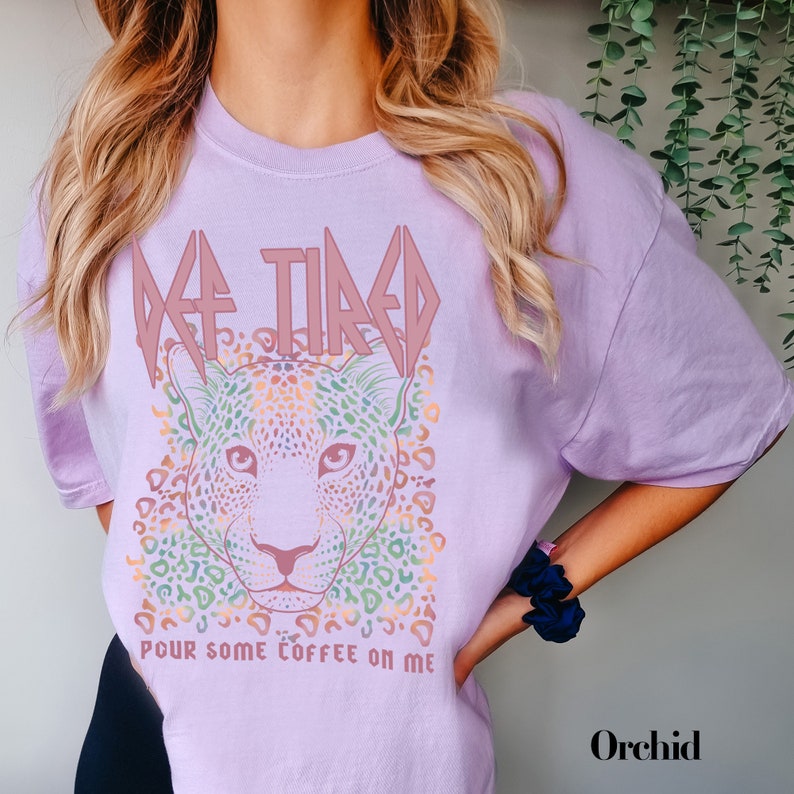 Puede incluir: Una camiseta lavanda con un estampado de leopardo colorido y el texto "DE-TIRED POUR SOME COFFEE ON ME" en rosa.