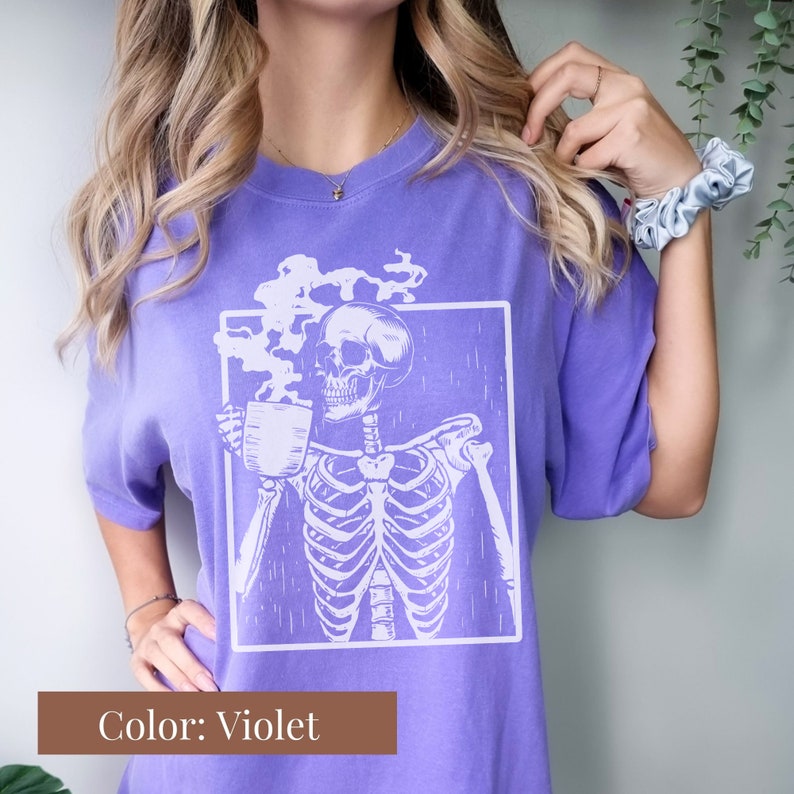 May include: Violet-colored t-shirt featuring a white graphic of a skeleton holding a coffee mug, with steam rising from the cup. The skeleton is inside a white square. The text "Color: Violet" is in a brown rectangle at the bottom.