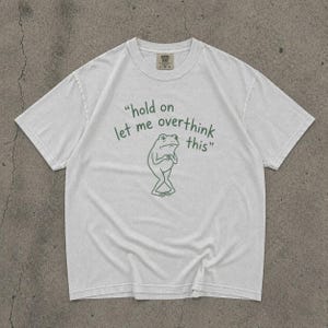 May include: A white t-shirt featuring a green frog illustration and the phrase "hold on let me overthink this." The shirt has a crew neck and short sleeves. The frog graphic shows a thoughtful pose.