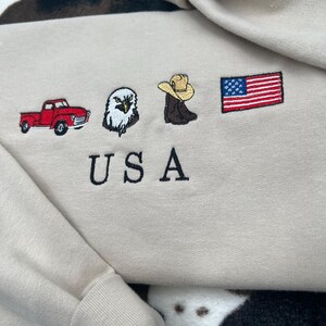 May include: Beige sweatshirt with embroidered images of a red pickup truck, a bald eagle, a cowboy boot with a hat, and an American flag. The word "USA" is embroidered below the images.
