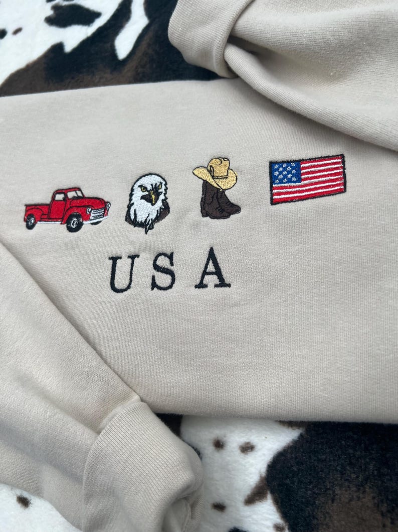 May include: Beige sweatshirt with embroidered images of a red pickup truck, a bald eagle, a cowboy boot with a hat, and an American flag. The word "USA" is embroidered below the images.