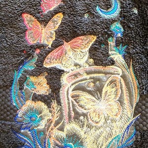 May include: Embroidered towel with a dark brown background featuring a crescent moon, stars, and multiple butterflies in shades of pink, yellow, and orange. The butterflies are surrounded by stylized flowers with blue, green, and white accents.