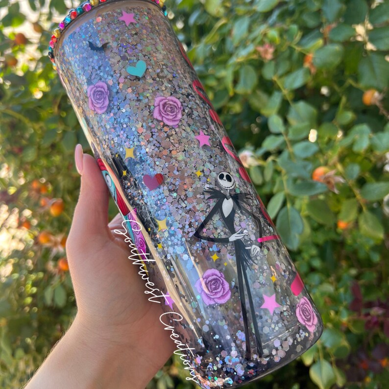 Puede incluir: Un vaso de pl&aacute;stico transparente con una tapa de pl&aacute;stico transparente. El vaso est&aacute; cubierto de purpurina negra y plateada y presenta un dise&ntilde;o de Jack Skellington de Pesadilla antes de Navidad, junto con corazones, estrellas y rosas. El vaso est&aacute; decorado con un borde de purpurina plateada y una base de purpurina plateada. El texto "Southwest Creations" est&aacute; impreso en el lateral del vaso.
