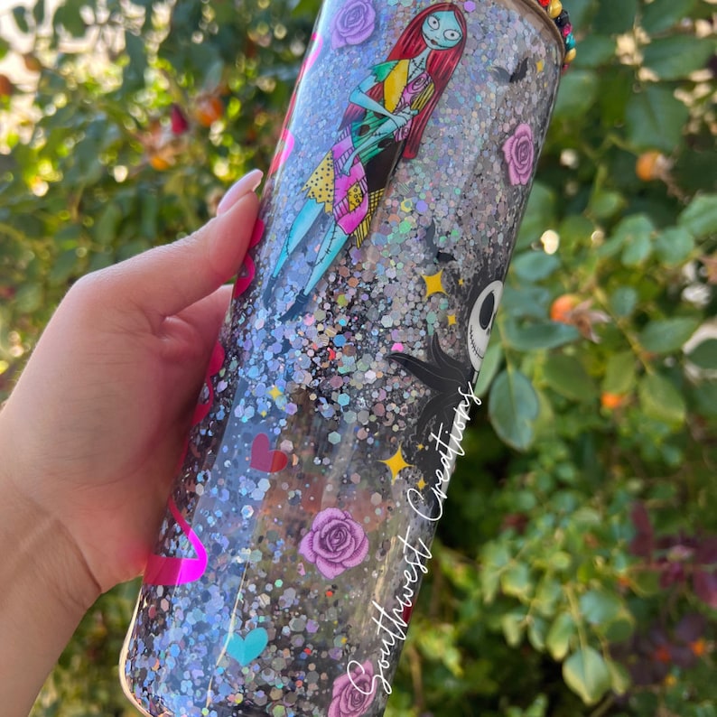 Puede incluir: Un vaso transparente con fondo de purpurina plateada y detalles de purpurina de colores. El vaso presenta un gr&aacute;fico de Jack Skellington y Sally de Pesadilla antes de Navidad. El vaso tambi&eacute;n tiene el texto "Southwest Creations" en el lateral.