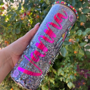 Puede incluir: Un vaso transparente con una base de purpurina negra y una parte superior de purpurina rosa. El vaso tiene un dise&ntilde;o de vinilo rosa que dice "Yanna" y un dise&ntilde;o de vinilo negro que dice "Southwest Creations". El vaso tiene un borde de cuentas de colores.