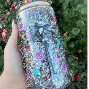 Puede incluir: Un vaso de vidrio transparente con fondo de purpurina plateada. El vaso presenta una imagen en blanco y negro de Jack Skellington de Pesadilla Antes de Navidad. El vaso tambi&eacute;n tiene corazones y estrellas de purpurina roja, rosa, azul y verde. El texto "Southwest Creations" est&aacute; en el lateral del vaso.