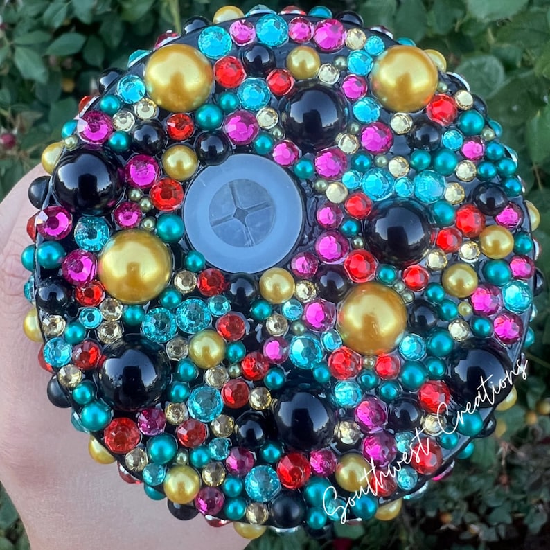 Puede incluir: Un tap&oacute;n de botella redondo negro con un centro transparente y un dise&ntilde;o de cuentas de colores, incluyendo oro, rojo, rosa, azul y verde.